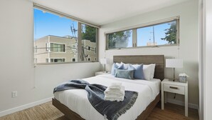 A bedroom with a large window, a bed with white bedding, two bedside tables with lamps, and a view of buildings outside. - Spacious Townhome in an Unbeatable Location (Seattle)