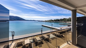 Uma varanda com vista para um mar calmo, um cais e montanhas ao longe.