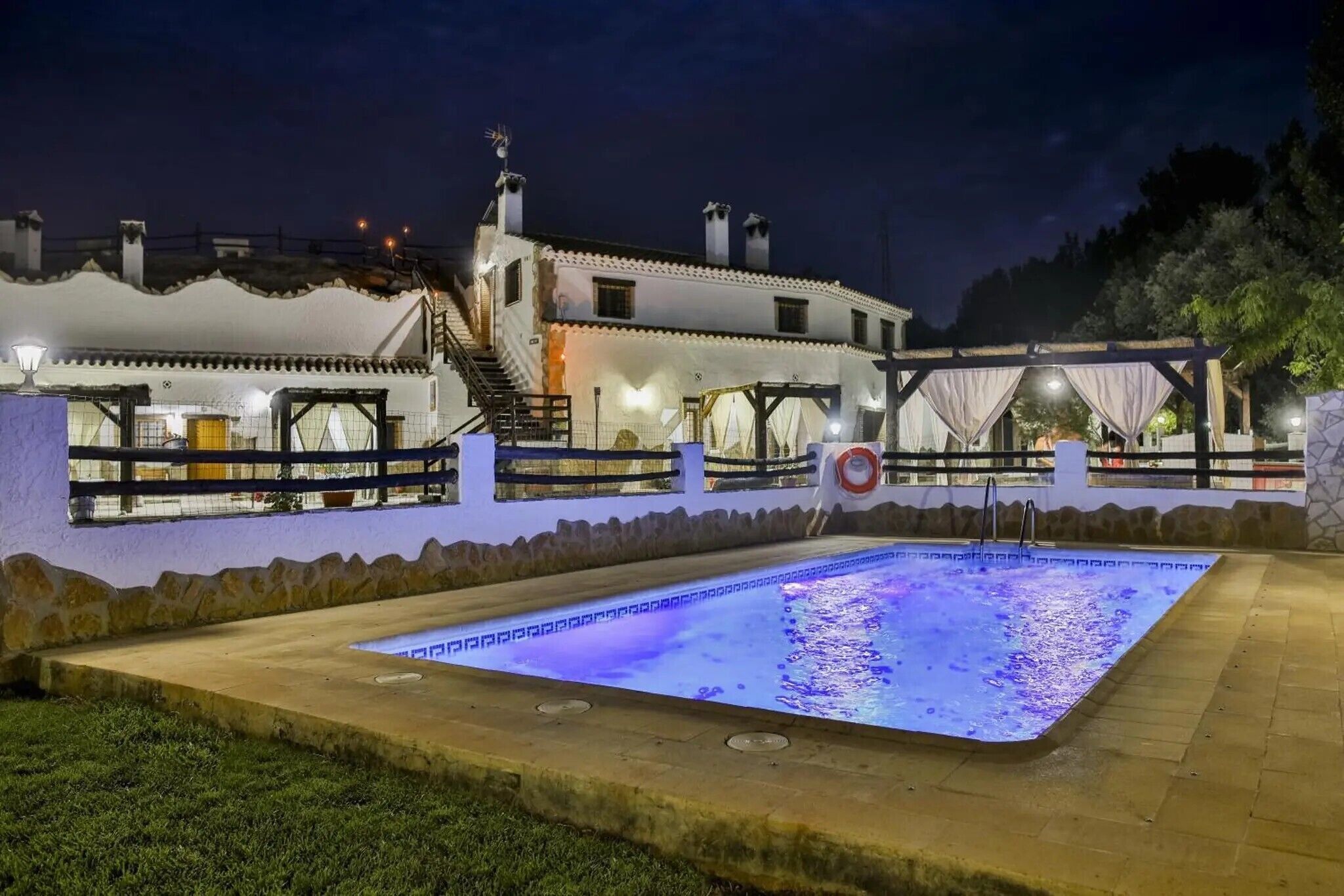 Un área de piscina con borde de piedra, una zona de estar cubierta y un edificio con varias ventanas y una escalera.