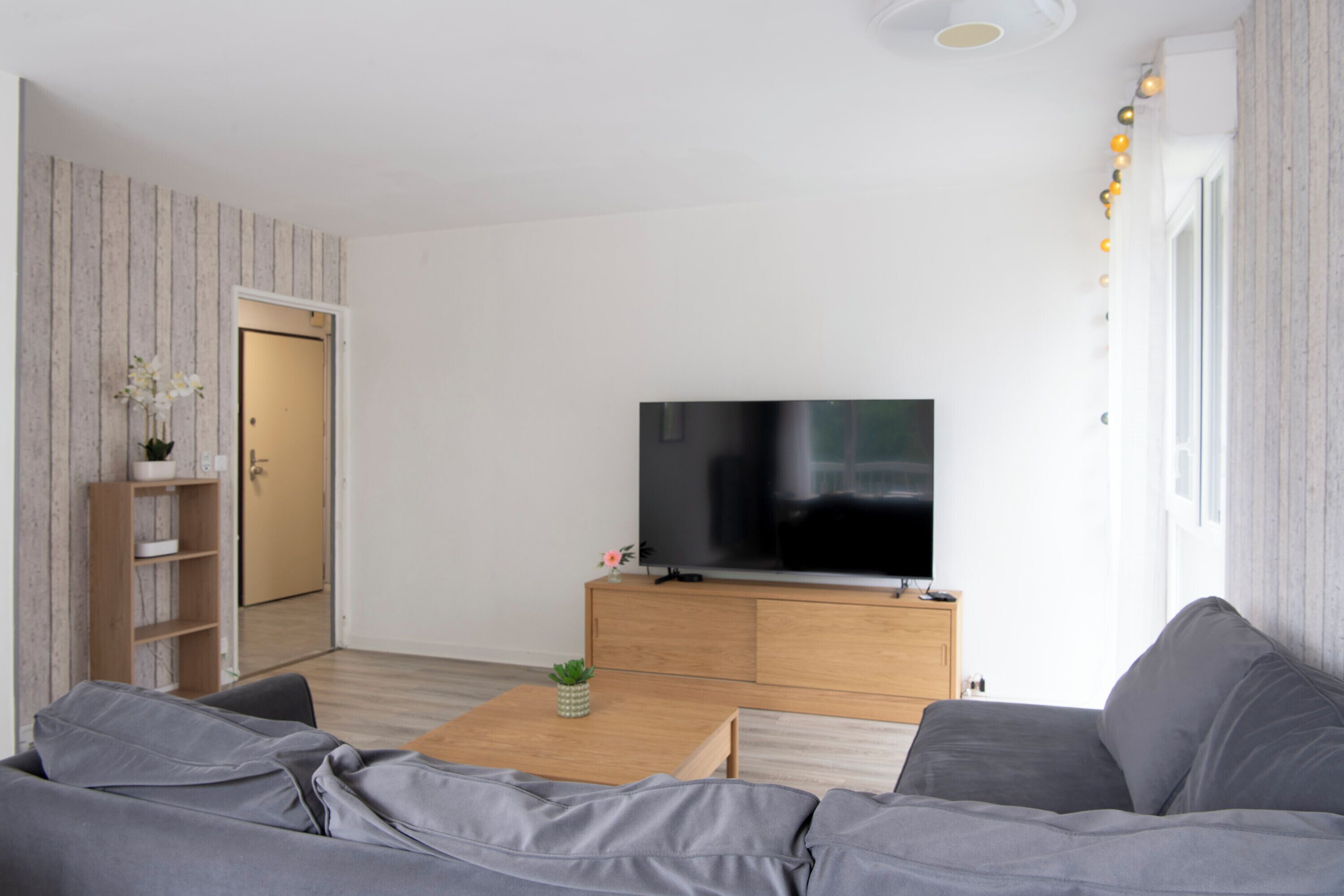 A modern living room with a flat-screen TV, grey sofa, wooden coffee table, and a bookshelf.
