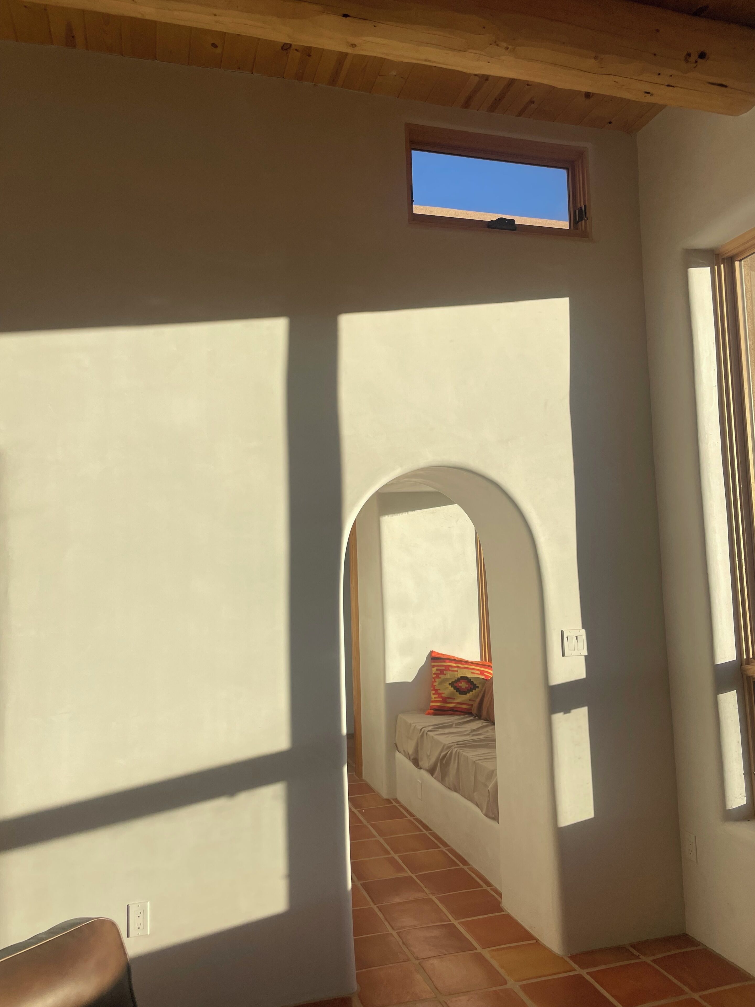 Una habitación con techo inclinado, un ventanal pequeño, una cama con almohada colorida y suelo de baldosas.