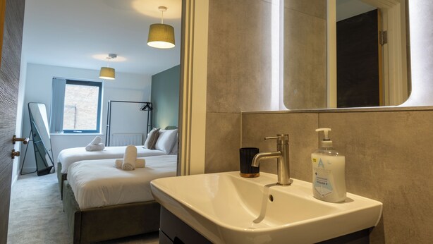 A hotel room with a large bed, a bedside table, a mirror, a sink with a soap dispenser, and a window. - Modern York Apartment - Free Parking - Sleeps 7 (York)