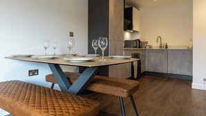 A modern dining area with a table set for four, a kitchen in the background, and a bench with a quilted seat. - Modern York Apartment - Free Parking - Sleeps 7 (York)