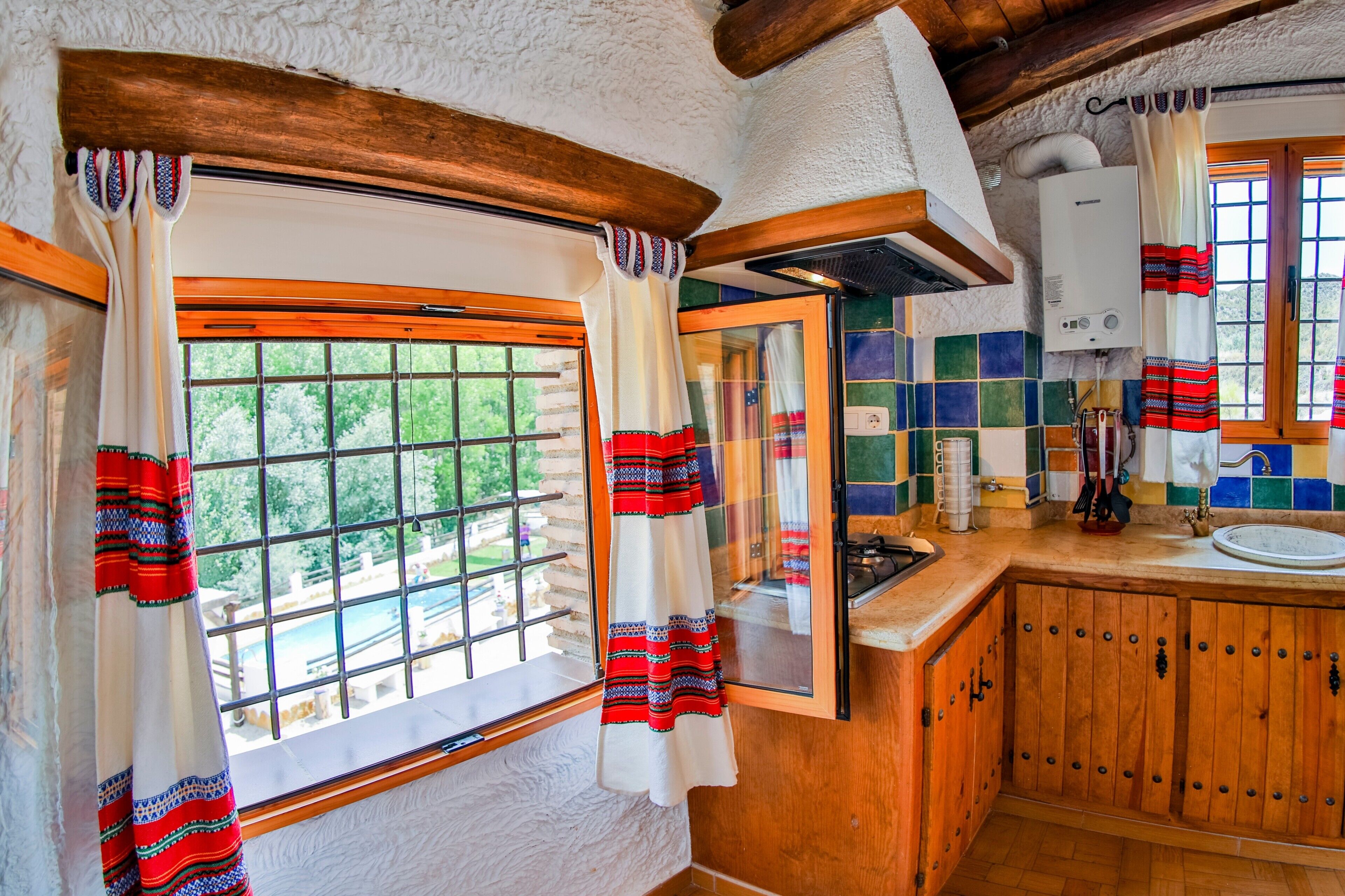 Une cuisine rustique avec des meubles en bois, une fenêtre agrémentée de rideaux et une vue sur la verdure à l’extérieur.