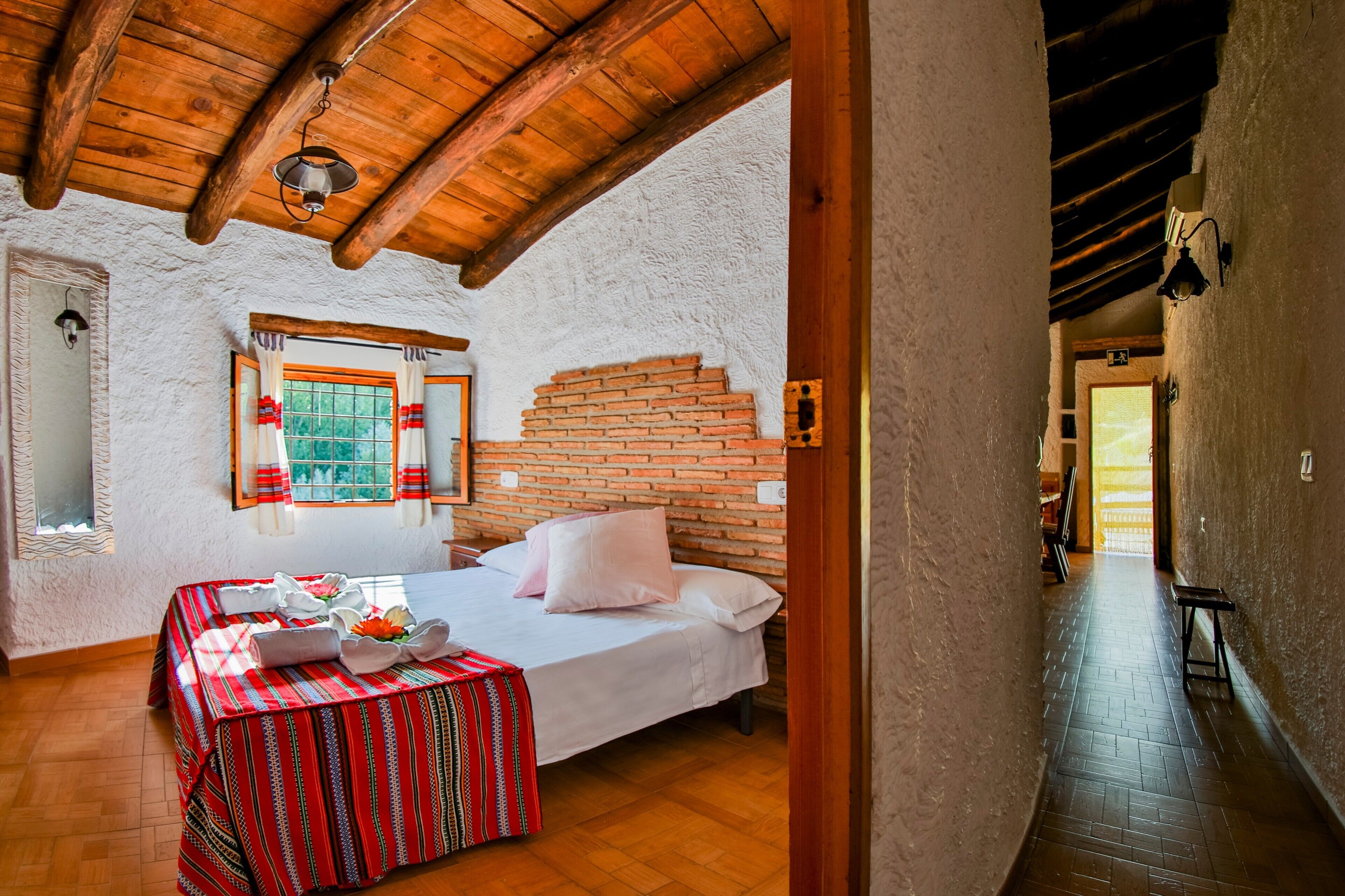 Una habitación con una cama, una mesa con mantel rojo, una pared de ladrillo y un techo de madera.