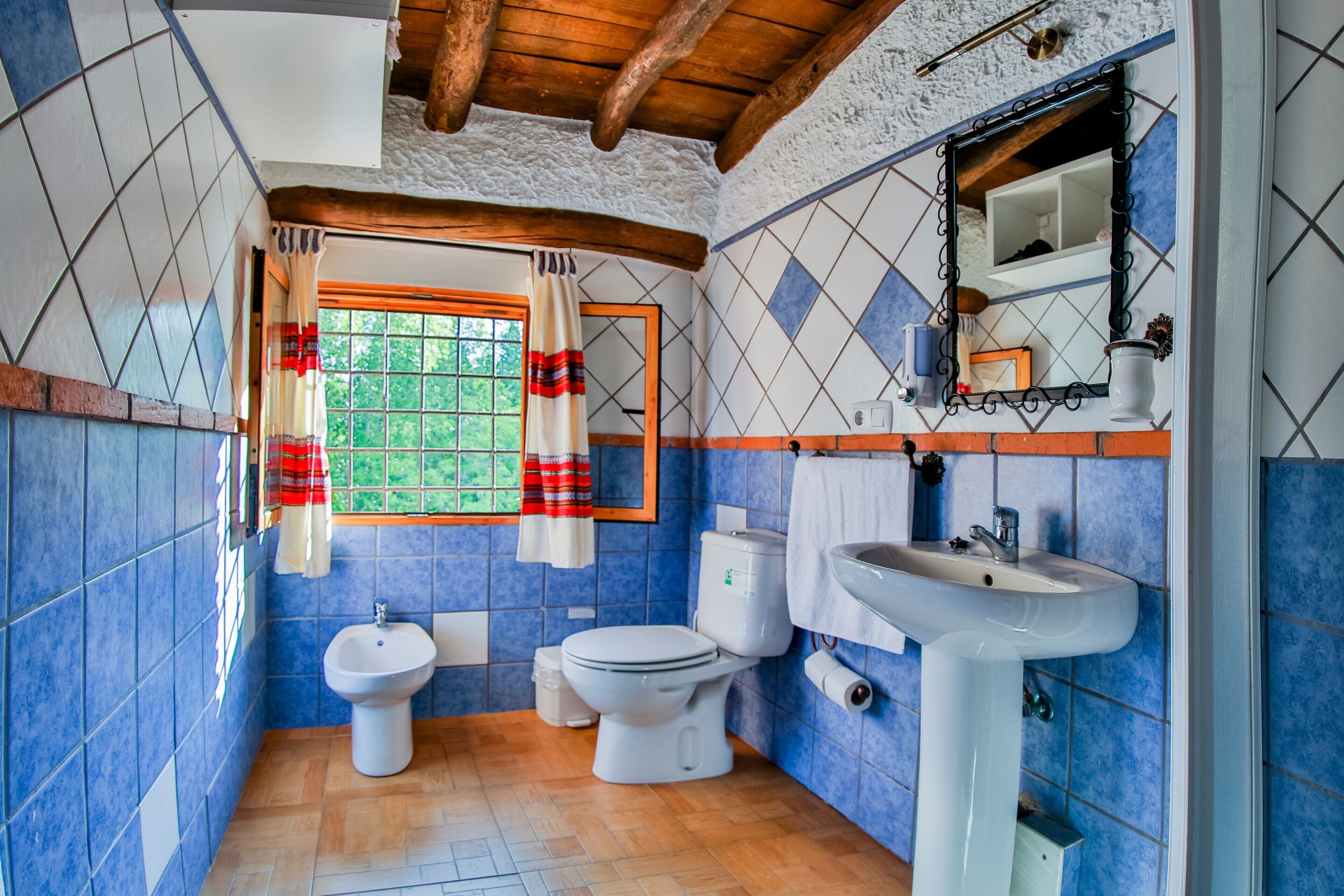 Un baño con azulejos azules, techo de madera, ventana con cortinas, un lavabo y dos inodoros.