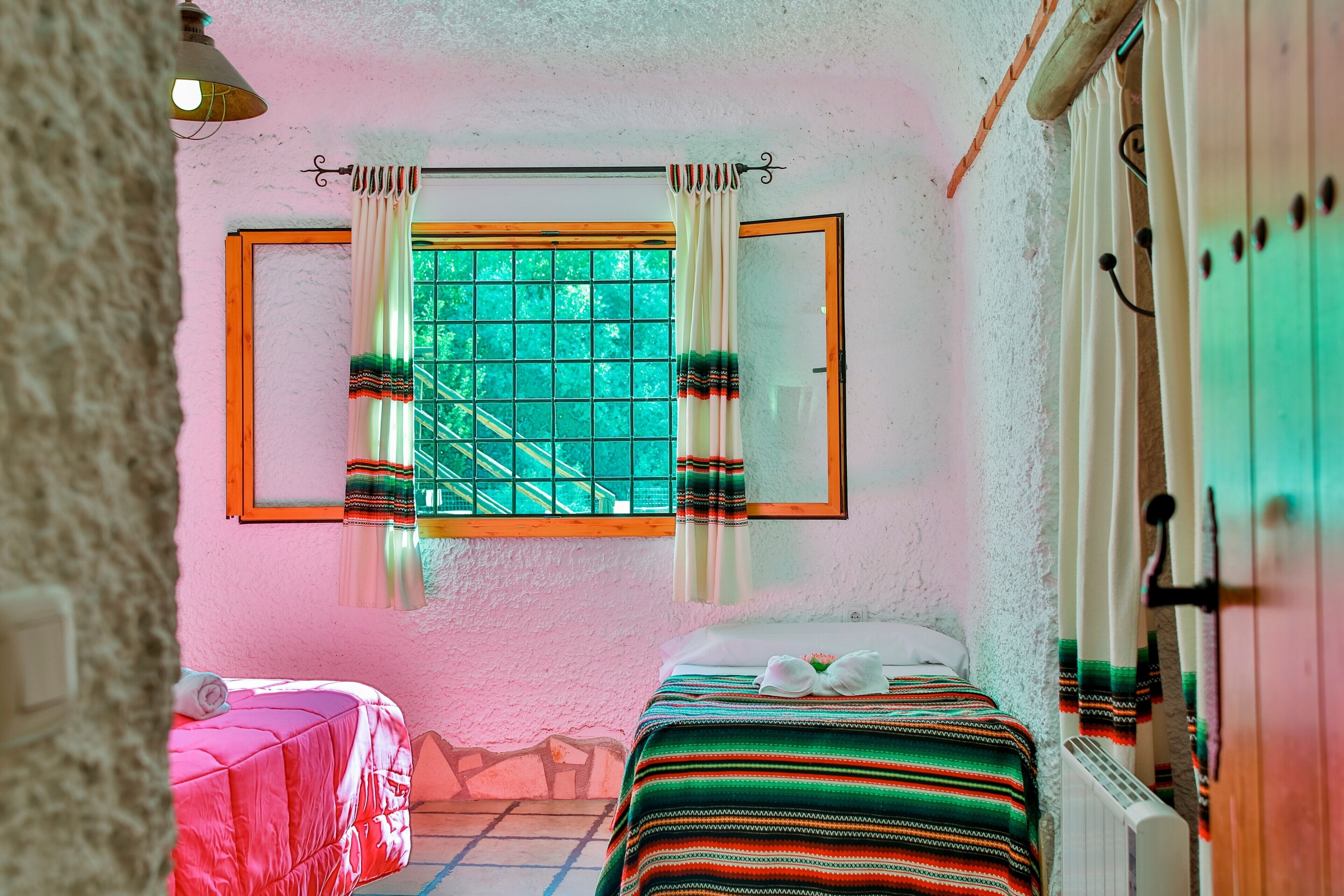A room with two beds, a window with curtains, a radiator, and a wall-mounted light fixture.