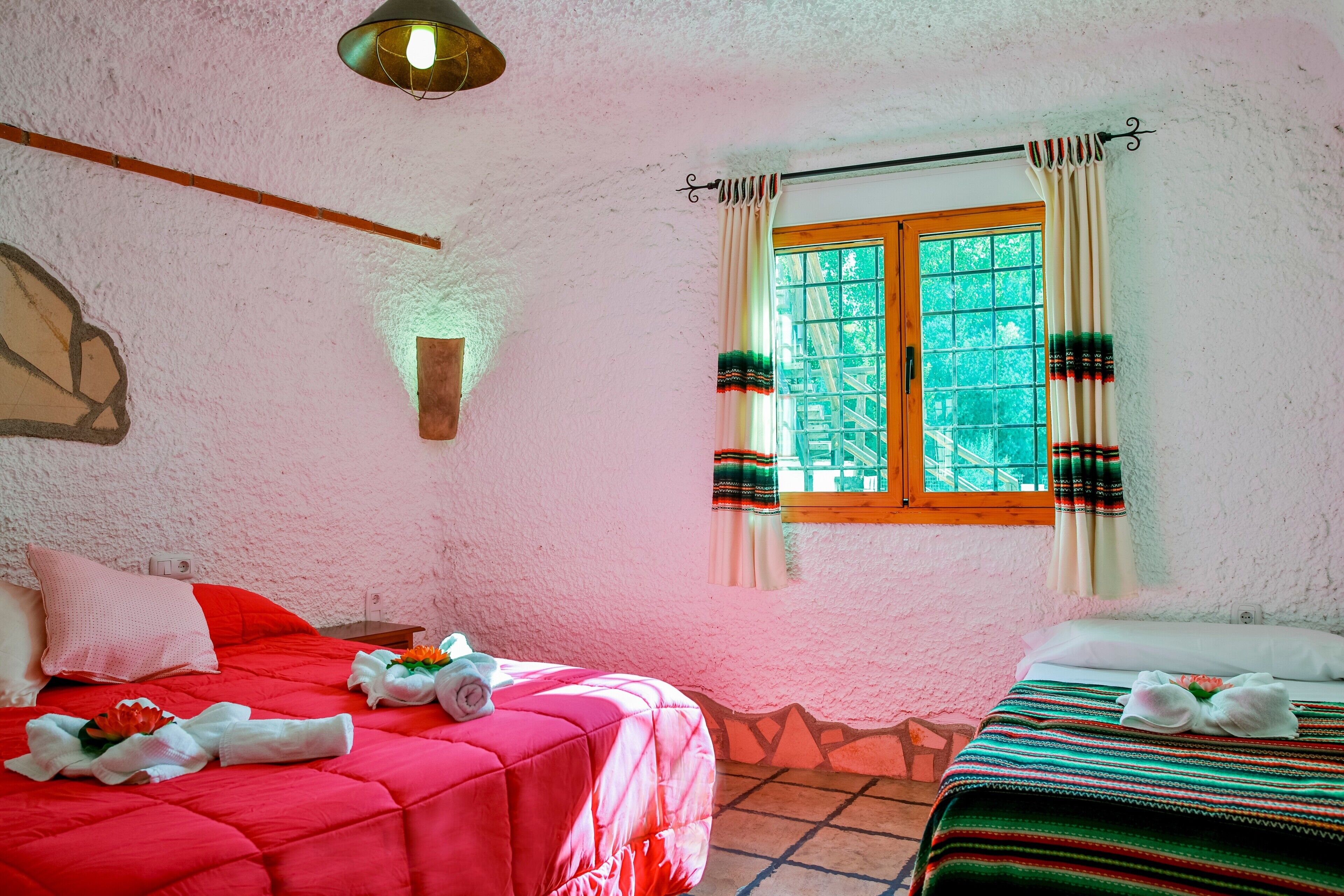 A room with two beds, a window with curtains, a wall-mounted light fixture, and a textured wall.