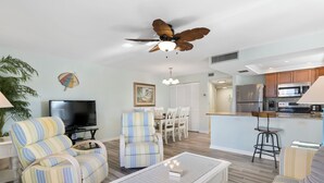 A living room with a TV, striped chairs, a ceiling fan, and a dining area with a table and chairs. - Gulf Gate Resort (St. Pete Beach)