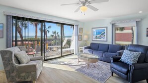A living room with a large sliding glass door leading to a balcony, a ceiling fan, and a view of palm trees and a beach. - Gulf Gate Resort (St. Pete Beach)