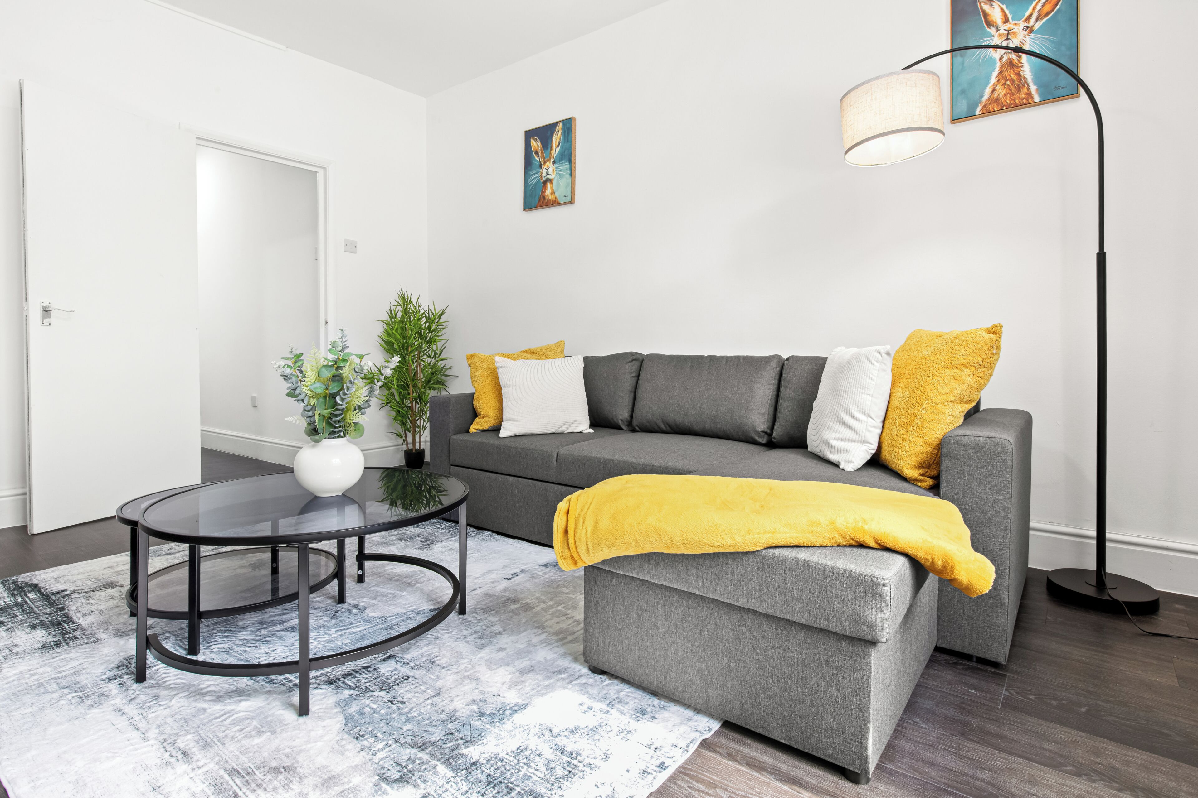 Ein modernes Wohnzimmer mit einem grauen Sofa, gelben Kissen, einem runden Couchtisch, einer Stehlampe und einem Bild an der Wand.