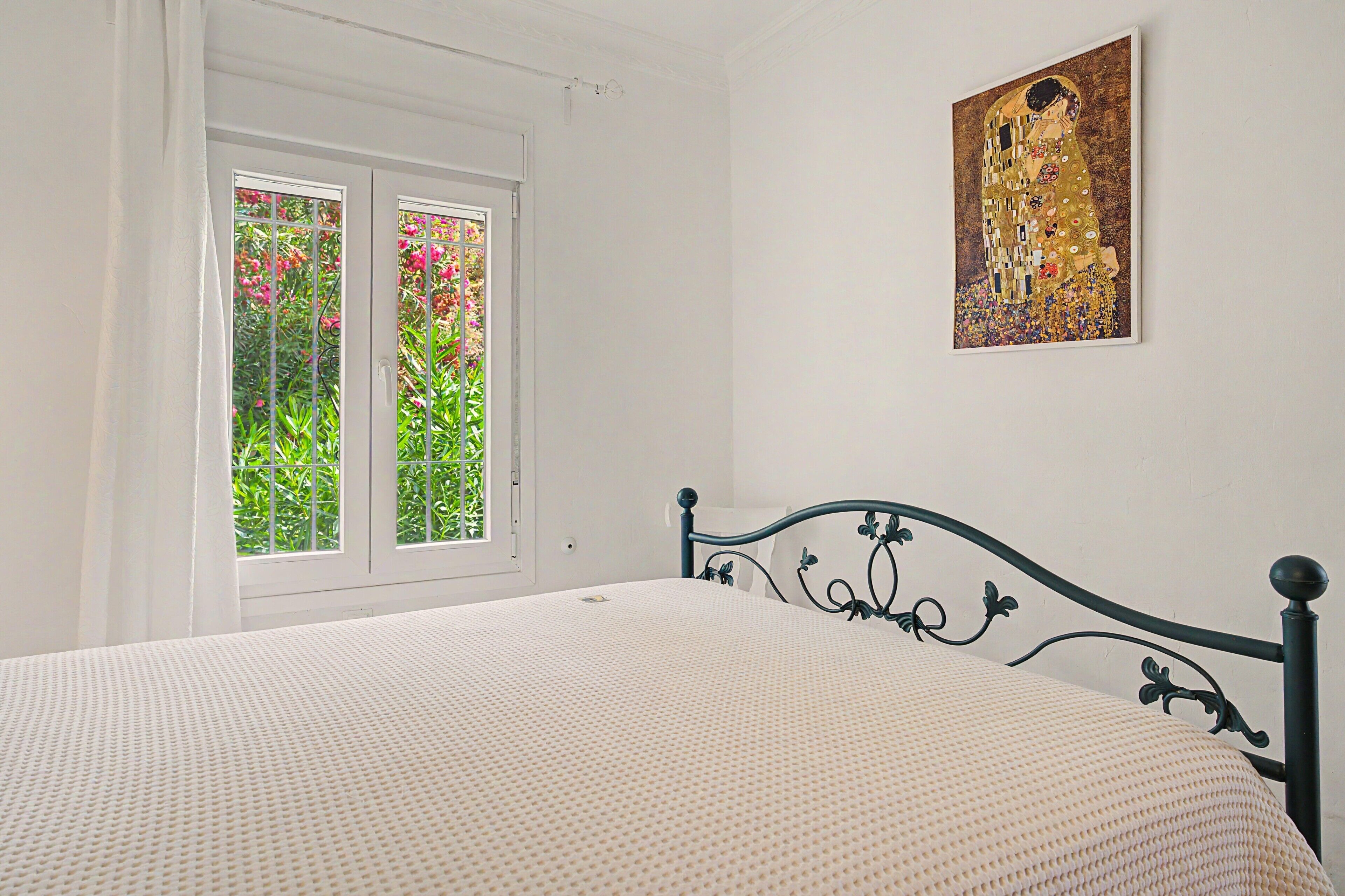 A bedroom with a bed, a window with a view of greenery, and a painting on the wall.
