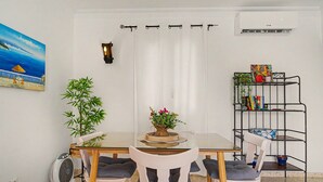 Un coin repas avec une table en verre, des chaises blanches, une plante en pot, un ventilateur fixé au mur et une bibliothèque.