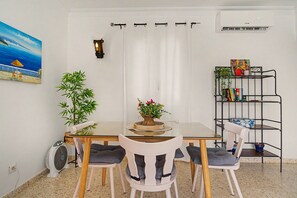 A dining area with a glass table, white chairs, a potted plant, a wall-mounted fan, and a bookshelf.