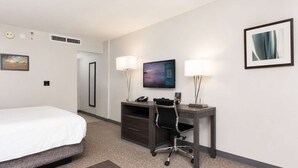Habitación de hotel con cama, escritorio, silla, dos lámparas, televisor y un cuadro en la pared.