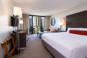 A hotel room with a large bed, a desk with a chair, a sofa, a TV, and a balcony with a view of tall buildings. - The Aqua Palms | Near Waikiki Beach + Free Parking (Honolulu)