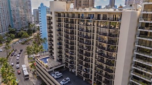 Un hotel de varios pisos con piscina en la azotea, rodeado de palmeras y otros edificios.