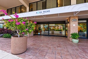 L’extérieur de l’hôtel Aqua Palms, avec son grand hall d’entrée, ses plantes en pot et une enseigne indiquant l’adresse.
