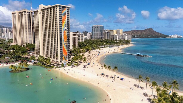 Un paisaje urbano costero con un hotel destacado que presenta un diseño de arcoíris vertical, playa, palmeras y un cielo azul despejado.