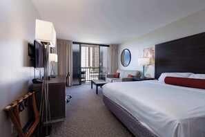 Ein Hotelzimmer mit einem großen Bett, einem Schreibtisch, einem Sessel, einem Fernseher und einem Balkon mit Vorhängen.