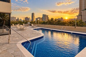 Una piscina con sillones de descanso y vista a la silueta urbana al atardecer.