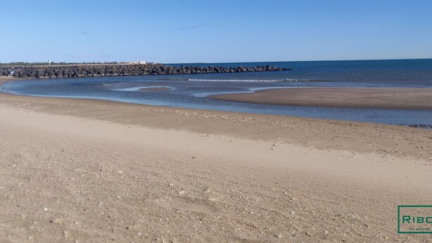 Una playa arenosa con un rompeolas rocoso y mar tranquilo.