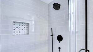 Zona de ducha con paredes de azulejos blancos, puerta de cristal y cabezal de ducha negro.