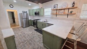 Una cocina con gabinetes verdes, un refrigerador de acero inoxidable, un microondas y una estufa. El piso presenta un patrón de espiga y una encimera blanca.