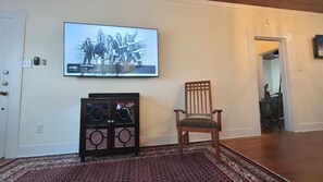 Ein Wohnzimmer mit an der Wand montiertem Fernseher, der ein Musikvideo zeigt, einem Holzstuhl mit gemustertem Kissen und einer dekorativen Vitrine mit Rundungen.