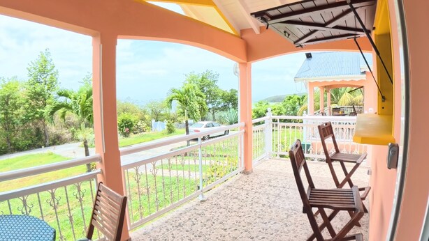 A covered balcony with a tiled floor, a chair, a table, and a view of a road and greenery. - Creole bungalow with views of the ocean and tropical scenery – Le Tapeur, Guadeloupe (Pointe-Noire)