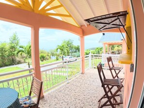 A covered balcony with a tiled floor, a chair, a table, and a view of a road and greenery. - Creole bungalow with views of the ocean and tropical scenery – Le Tapeur, Guadeloupe (Pointe-Noire)