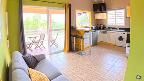 A living room with a sofa, a dining area with a table and chairs, a kitchen with appliances, and a sliding glass door leading to an outdoor area. - Creole bungalow with views of the ocean and tropical scenery – Le Tapeur, Guadeloupe (Pointe-Noire)