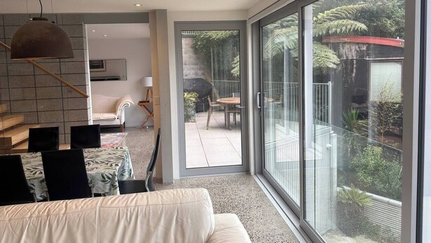 A modern living room with a sofa, dining table, chairs, and a large sliding glass door leading to a balcony with outdoor furniture and greenery. - Stunning beachside family retreat (Wellington)