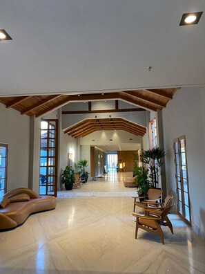 Un vestíbulo amplio con techo alto, vigas de madera y una escalera central. Varias zonas de estar con sillones y plantas en macetas.