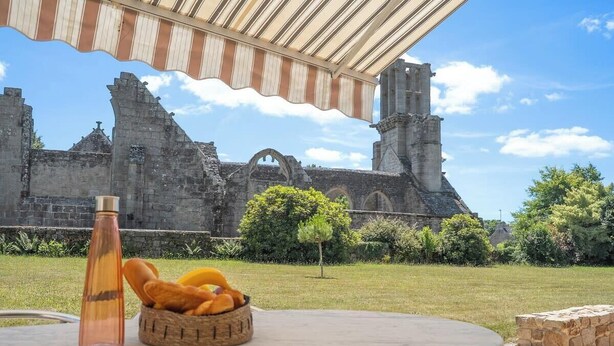 A table with a bottle and a basket of bread, set against a backdrop of ancient ruins under a striped awning. - Maison en pierre dans un hameau privé (Pont-l'Abbé)
