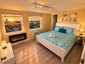 A bedroom with a bed, two windows with city views, a fireplace, and a nightstand with a lamp. - CasaViva Sky · Spacious 3BR w/ City Views, Firepit & Backyard, walk everywhere (San Diego)
