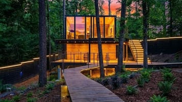 A modern cabin with a glass facade, surrounded by a wooden deck and a forested area.