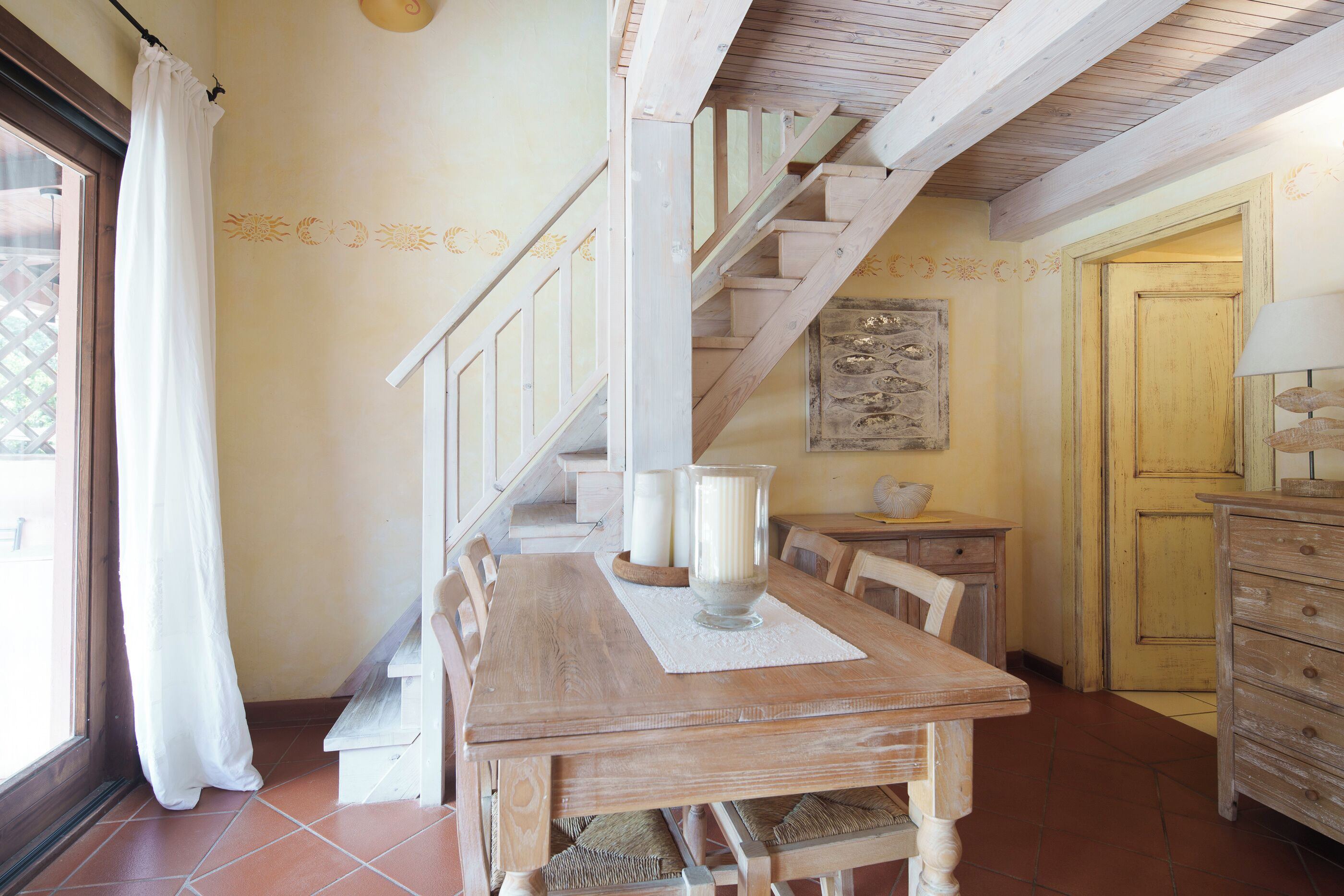 Une table à manger en bois avec des chaises, un escalier, un luminaire fixé au mur et une porte.