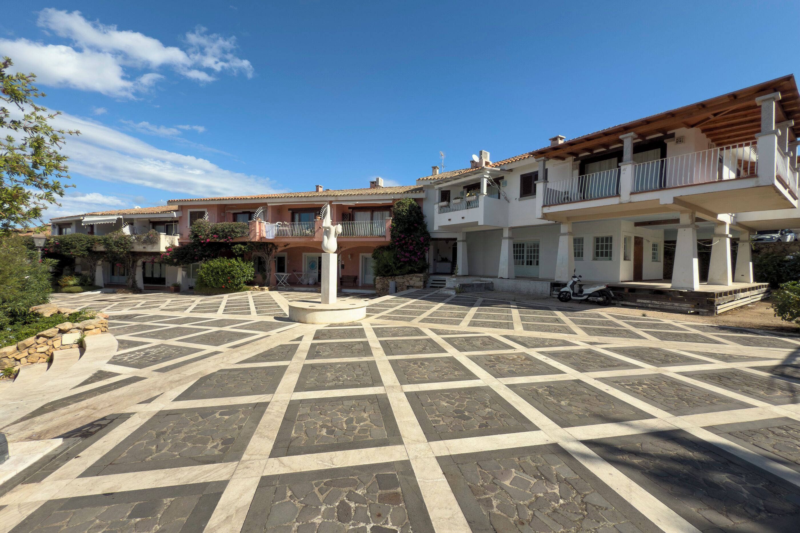 Un cortile con pavimento in piastrelle geometriche, una fontana al centro e edifici a più piani con balconi.