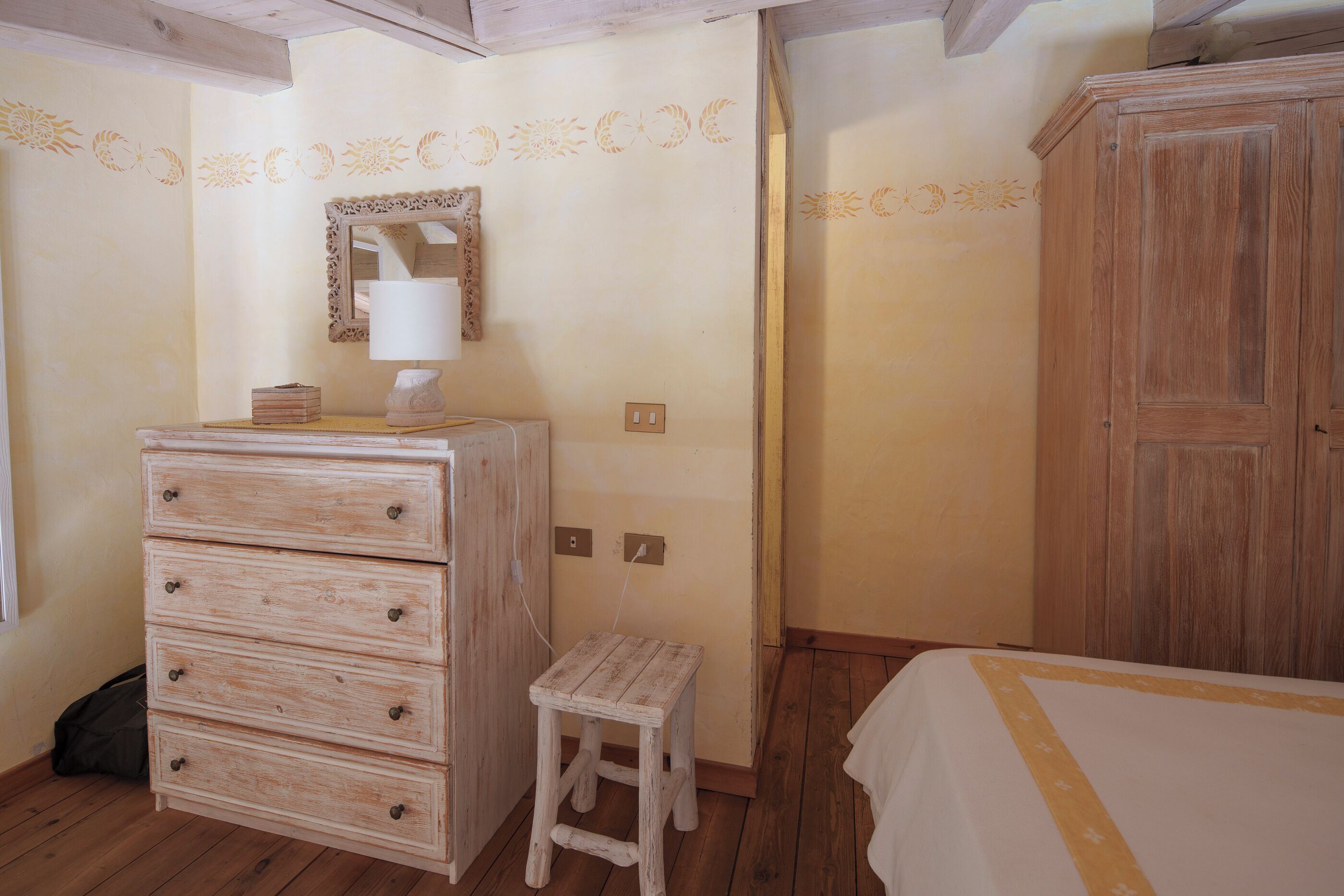 Una camera da letto con un comò in legno, uno sgabello, un letto con copripiumino bianco e un armadio in legno.