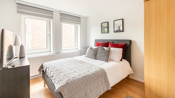 Un dormitorio con cama, mesitas de noche, armario y dos ventanas con persianas.