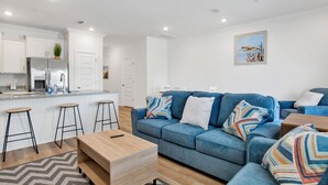 A modern living room with a blue sofa, wooden coffee table, and a kitchen area with white cabinets and a stainless steel refrigerator. - Brand New 3-bedroom townhome with AC in the heart of Orange Beach (Orange Beach)