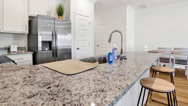 A kitchen with white cabinets, a stainless steel refrigerator, a granite countertop, a sink, and two wooden bar stools. - Brand New 3-bedroom townhome with AC in the heart of Orange Beach (Orange Beach)