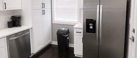 A modern kitchen with white cabinets, a stainless steel refrigerator, a dishwasher, and a window with blinds.