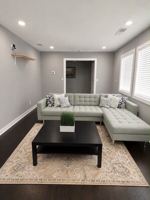 A living room with a sectional sofa, a coffee table, a patterned rug, a wall-mounted shelf, and a door leading to another room.