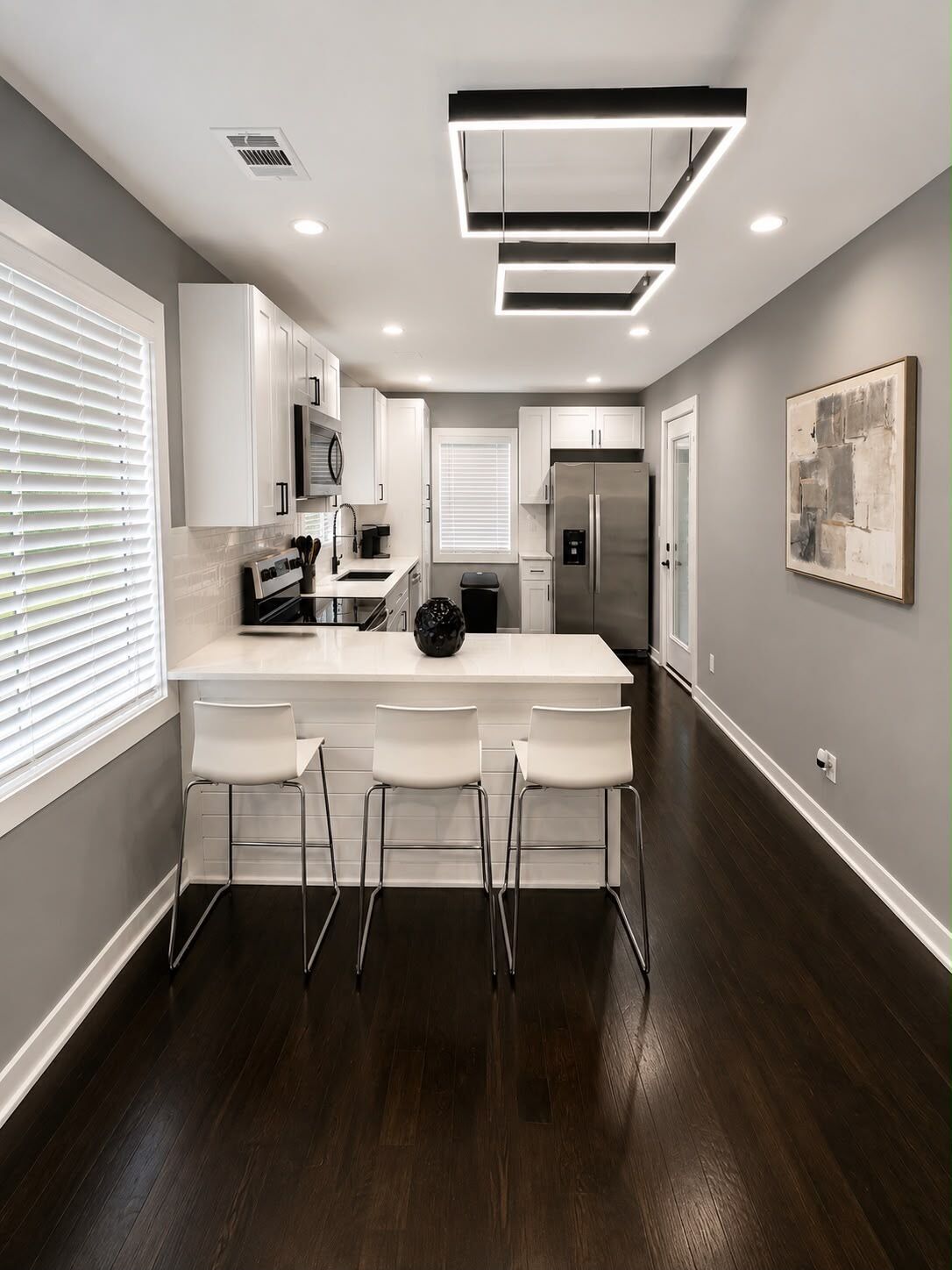 A modern kitchen with white cabinetry, a central island with bar stools, a stainless steel refrigerator, and a contemporary light fixture.