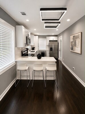 A modern kitchen with white cabinetry, a central island with bar stools, a stainless steel refrigerator, and a contemporary light fixture. - Cozy Modern Escape Near Downtown ATL (Atlanta)