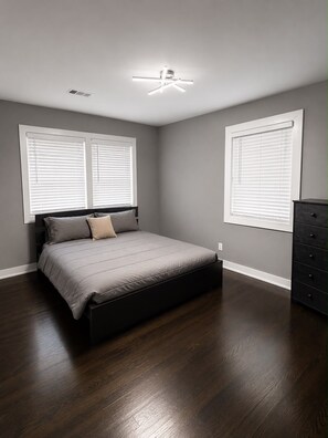 A bedroom with a bed, a dresser, and two windows with blinds.