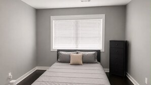 A bedroom with a bed, a window with blinds, a nightstand, and a ceiling light fixture.