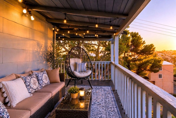 A balcony with a sofa, a hanging chair, a small table with a plant, and string lights. - CasaViva Vista · Top-Floor 2BR with Private Porch, City Views, Walk to Eats (San Diego)